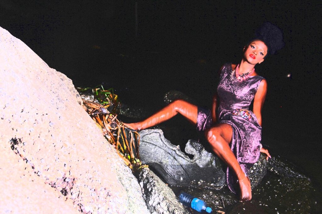 Ify Otuya sitting on a rock and surrounded by water in Couture Art Photograph.