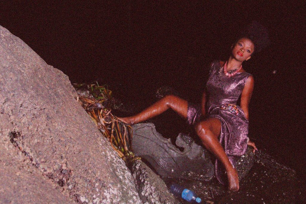 Model/ Singer Ify Otuya sitting on a rock and surrounded by water in Couture Art Photograph.